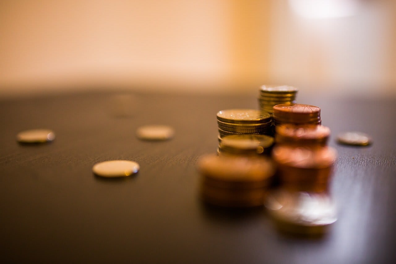 A close-up of various coins stacked on a dark surface, with some coins scattered around. The background is softly blurred.