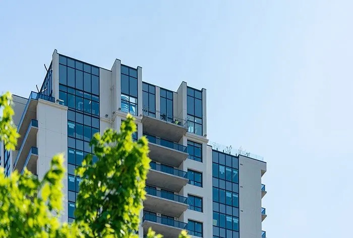 A modern high-rise building with glass windows and balconies, partially obscured by green foliage, against a clear blue sky.