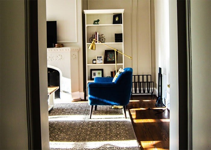A cozy living room featuring a blue chair, a bookshelf, and a fireplace, with sunlight streaming in.